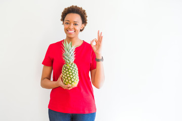 Young african american woman holding fresh healthy pineapple fruit doing ok sign with fingers, excellent symbol