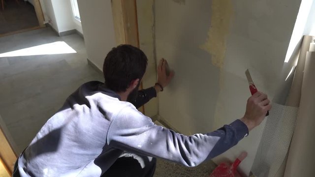 Young Caucasian Brown Haired Beard Man in Dark Purple Pullover and Track Pants is Measuring Fixing and Cutting with a Stanley Knife the Plaster Mesh Reinforcement on the Wall