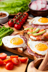 Huge healthy breakfast with sandwiches with scrambled eggs, sausage, salmon, arugula, curd cheese, avocado on a wooden board, sun-dried tomatoes, asparagus, radishes, greens on black table top view