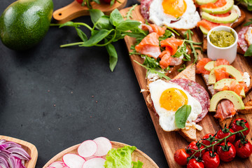 Huge healthy breakfast with sandwiches with scrambled eggs, sausage, salmon, arugula, curd cheese, avocado on a wooden board, sun-dried tomatoes, asparagus, radishes, greens on black table top view