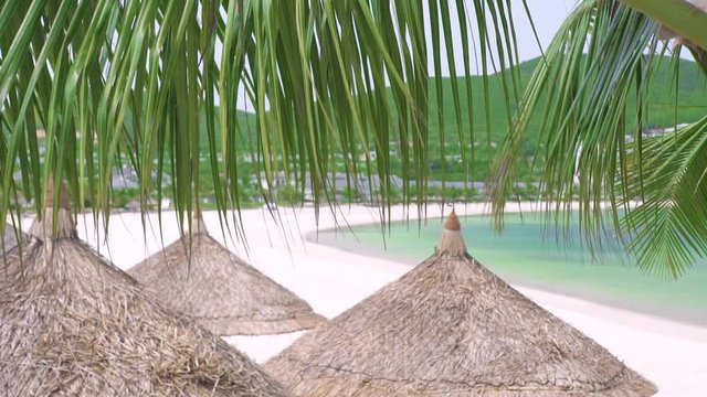 Straw umbrellas on sea beach on green mountains landscape and foliage of tropical palm trees. Paradise tropical beach, blue sea, green mountains and thatched umbrellas.