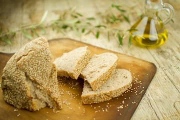 Close-up of a sliced loaf of homemade bread with sesame seeds, ampoule of extra virgin olive oil and an olive branch in selective focus on wooden table background