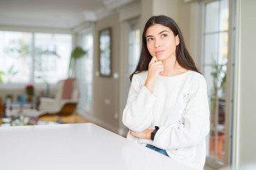 Obraz premium Young beautiful woman at home on white table with hand on chin thinking about question, pensive expression. Smiling with thoughtful face. Doubt concept.