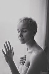 Beautiful young woman with short blond hair standing at the window. Black-and-white picture. Vertical picture.