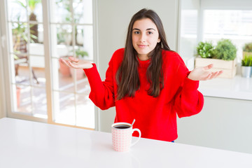 Beautiful young woman drinking a cup of black coffee clueless and confused expression with arms and...