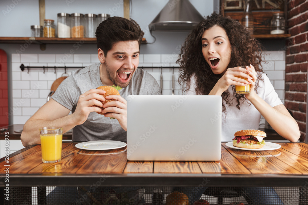 Canvas Prints Picture of young couple looking at laptop on table while eating hamburger in kitchen at home