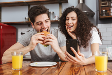 Picture of european using mobile phone while eating hamburger during breakfast in kitchen at home