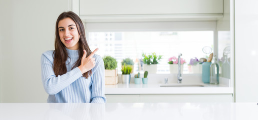 Wide angle picture of beautiful young woman sitting on white table at home cheerful with a smile of face pointing with hand and finger up to the side with happy and natural expression on face