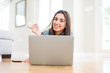 Fototapeta premium Beautiful young woman laying on the floor using laptop very happy pointing with hand and finger to the side