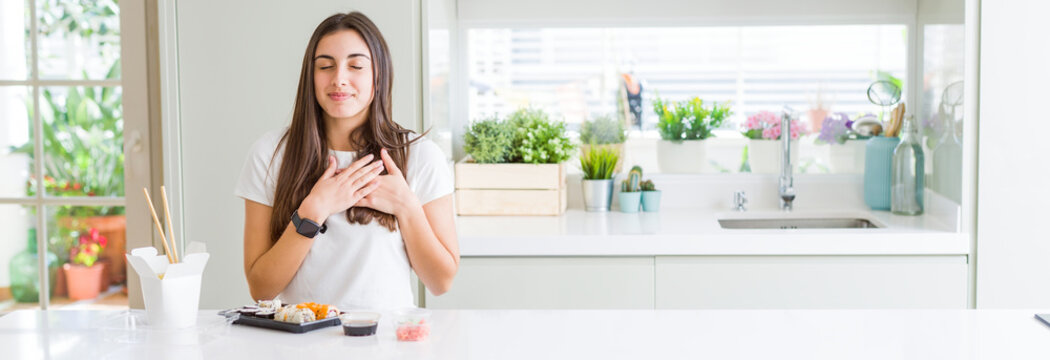 Wide angle picture of beautiful young woman eating asian sushi from delivery smiling with hands on chest with closed eyes and grateful gesture on face. Health concept.