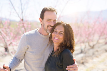 Naklejka premium Beautiful middle age senior couple smiling in love at romantic garden of peach trees with pink petals on a sunny day of spring