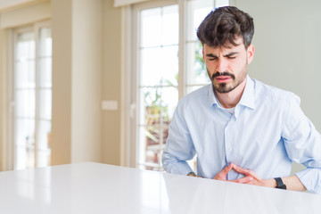 Fototapeta premium Young businesss man sitting on white table with hand on stomach because indigestion, painful illness feeling unwell. Ache concept.
