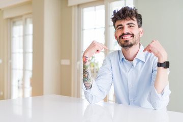 Young businesss man sitting on white table looking confident with smile on face, pointing oneself with fingers proud and happy.