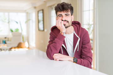 Young man wearing casual sweatshirt sitting on white table looking stressed and nervous with hands on mouth biting nails. Anxiety problem.