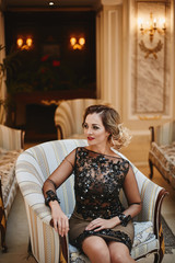 Obraz na płótnie Canvas Young and sexy businesswoman in a stylish cocktail dress sits in a vintage armchair at luxury interior