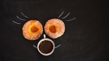 Two donuts with apricot jam lie and a Cup of coffee on a black Board. Chalk painted eyelashes and smile. The face of an unusual character from bakery products. A baby with a pacifier.