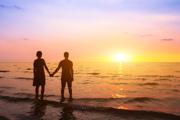 Romantic couple on the beach at sunset watching horizon, honeymoon vacation holidays at sea destination, silhouette of two lovers holding hand