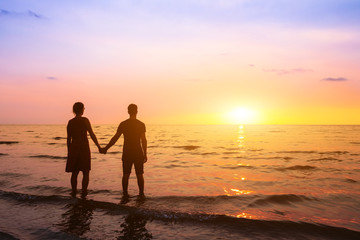 Fototapeta premium Romantic couple on the beach at sunset watching horizon, honeymoon vacation holidays at sea destination, silhouette of two lovers holding hand