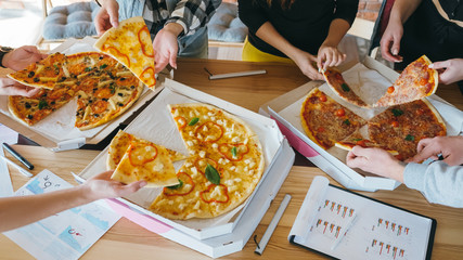 Millennials business lunch. Young team having break after successful work. Closeup of pizza in boxes on desk with charts.