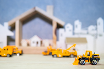 Miniature yellow tractor model with blurred house frame and city background on wooden table. Architecture and construction industry concept