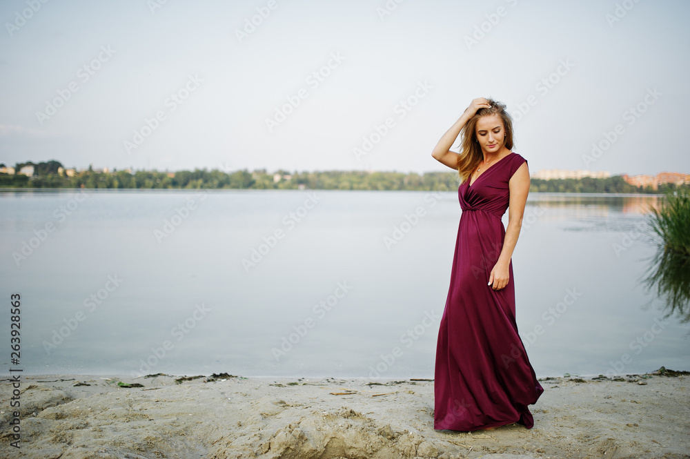 Canvas Prints Blonde sensual woman in red marsala dress posing against lake with reeds.