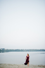 Blonde sensual barefoot woman in red marsala dress posing against lake on sand.