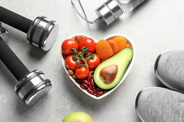 Heart shaped bowl with healthy products and sports equipment on grey background, flat lay