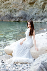 Fototapeta na wymiar Full length portrait of elegant bride on a rocky beach. Young beautiful brunette woman in long light grey chiffon wedding dress embroidered with beads sitting on a rock near the sea.