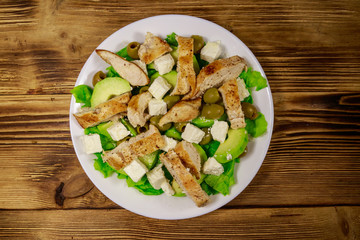 Tasty salad with fried chicken breast, green olives, feta cheese, avocado, lettuce and olive oil on wooden table. Top view