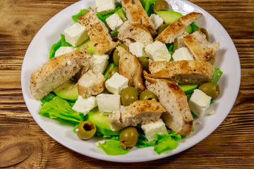 Tasty salad with fried chicken breast, green olives, feta cheese, avocado, lettuce and olive oil on wooden table