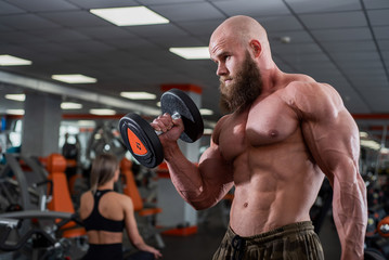 Fototapeta na wymiar Brutal, muscular, bald with a beard athlete in the gym. Lifts heavy weights, trains the biceps. Beautiful chest muscles