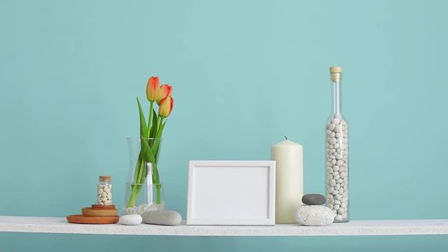 Modern room decoration with picture frame mockup. Shelf against turquoise wall with decorative candle, glass and rocks. Hand putting Tulips in vase.