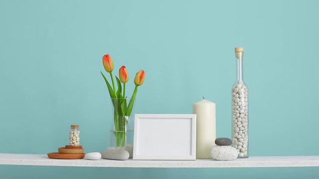 Modern room decoration with picture frame mockup. Shelf against turquoise wall with decorative candle, glass and rocks. Hand putting Tulips in vase.