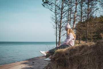 Naklejka na ściany i meble Young adult woman in pink hat sitting alone on the bluffs, looking of sea, freedom concept, peaceful atmosphere, meditation, copy space, Latvia, Jurkalne
