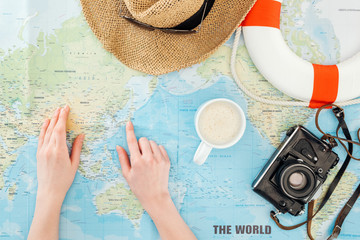 Fototapeta na wymiar Cropped view of woman with cup of cappuccino, film camera, sunglasses, lifebuoy and straw hat pointing with finger on world map