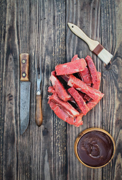Uncooked raw boneless beef ribs with bbq sauce, brush, vintage meat fork and butcher's knife over top a rustic wood table / background. Image shot from overhead view.
