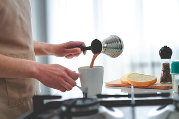 Woman in apron prepare hot tasty turkish coffee from cezve for breakfast at kitchen at home.
