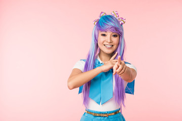 Smiling asian anime girl in purple wig pointing with finger at camera isolated on pink