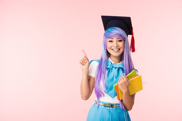 Smiling asian anime girl in academic cap holding books and pointing with finger isolated on pink