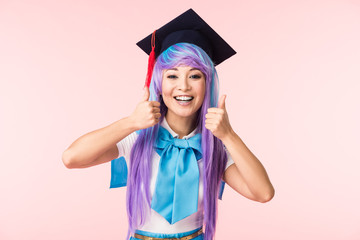 Front view of laughing asian anime girl in academic cap showing thumbs up isolated on pink