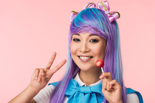 Happy asian anime girl holding lollipop and showing peace sign isolated on pink