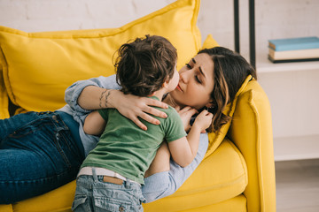 Obraz premium Little boy kissing tired mother lying on yellow sofa