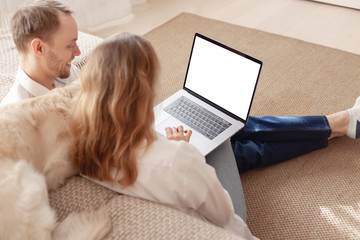 Rear view of a happy young married couple with a big white dog watching laptop with white screen in search of a suitable travel agency for a summer holiday
