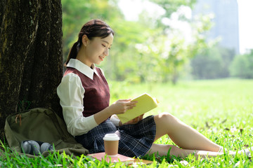 Naklejka na ściany i meble Portrait of beautiful smiling Asian woman sitting on green grass in park and relaxing while reading a book.