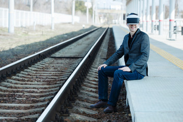 smiling young man extreme sits near the train tracks at the station wearing virtual reality glasses. Futuristic- digital technology concept. Virtual space. Vr headset and  glasses