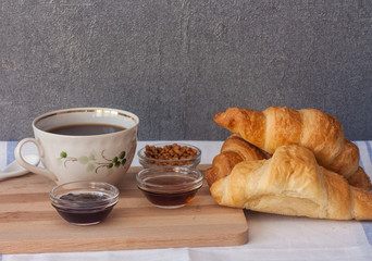 Three croissant, bowls with jam, syrup and nuts, a cup of coffee on a cutting wooden board on a light table on a background of gray wall. 