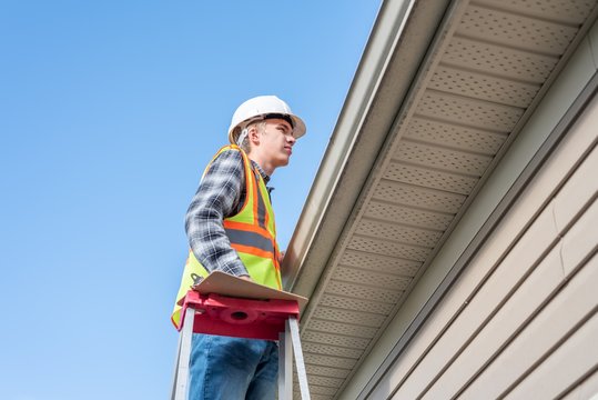 Home Inspector Standing On A Ladder And Providing An Inspection To The Roof Of A House.