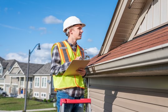 Home Inspector Standing On A Ladder And Providing An Inspection To The Roof Of A House.