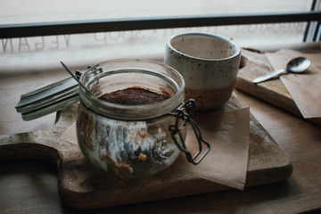 High angle view of tiramisu in glass cup with coffee cappuccino. In vegsn shop, local cozy cafe. Wooden background, candid style. Copy space