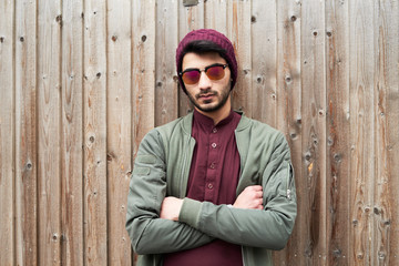Portrait of serious bearded man in purple knit hat and shirt with arms crossed wearing sunglasses over wooden background